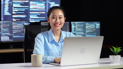 asian female programmer smiling to camera while writing code by a laptop using multiple monitors showing database on terminal window desktops in the office