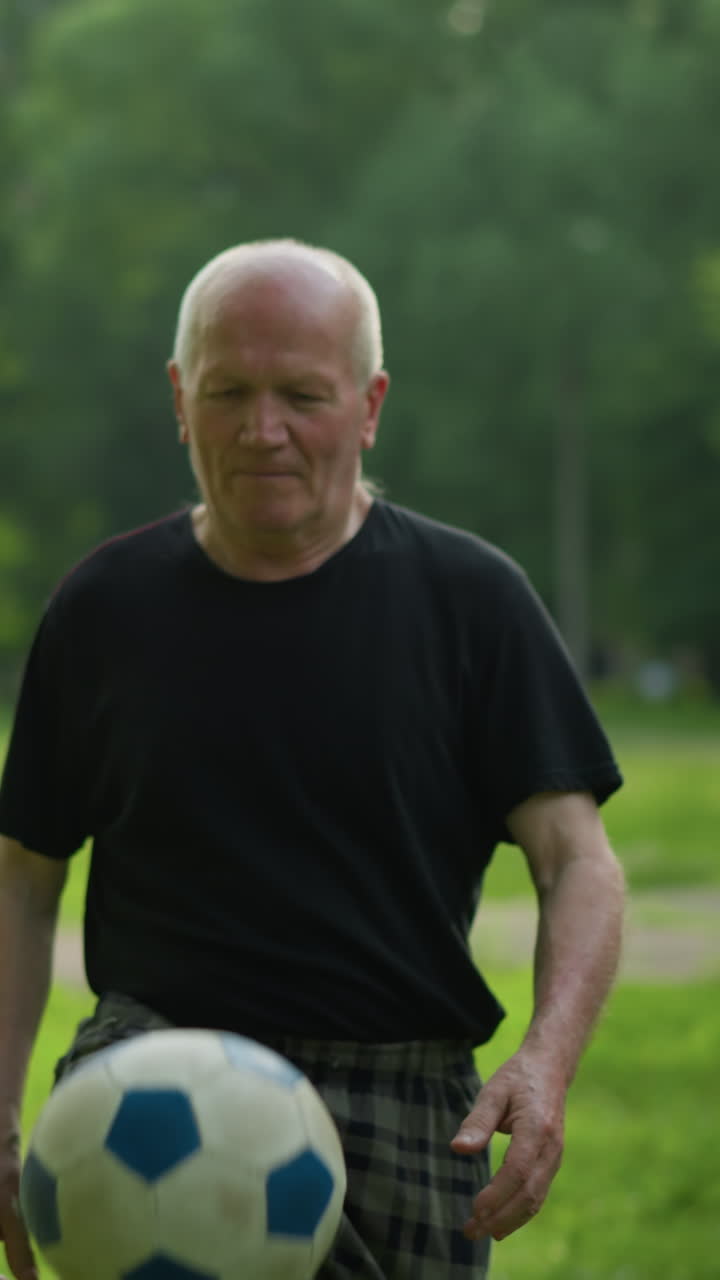 An elderly man skillfully taps a soccer ball up in a lush grassy field, surrounded by trees and a goalpost in the background, focused as he taps the ball and kicks it