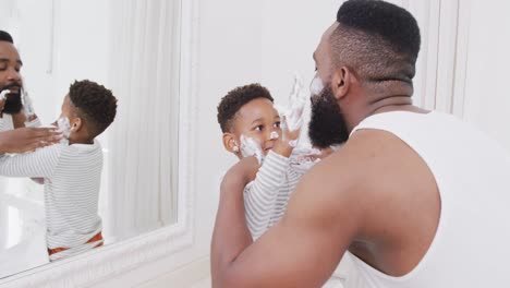 Happy-african-american-father-and-son-applying-shaving-cream-on-face-in-bathroom,-in-slow-motion