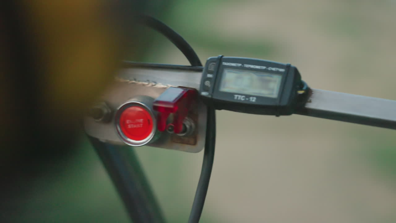 cockpit controls featuring bright red engine start button mounted on metal bar alongside digital tachometer display with wires and switch, blurred green rural landscape visible through windshield