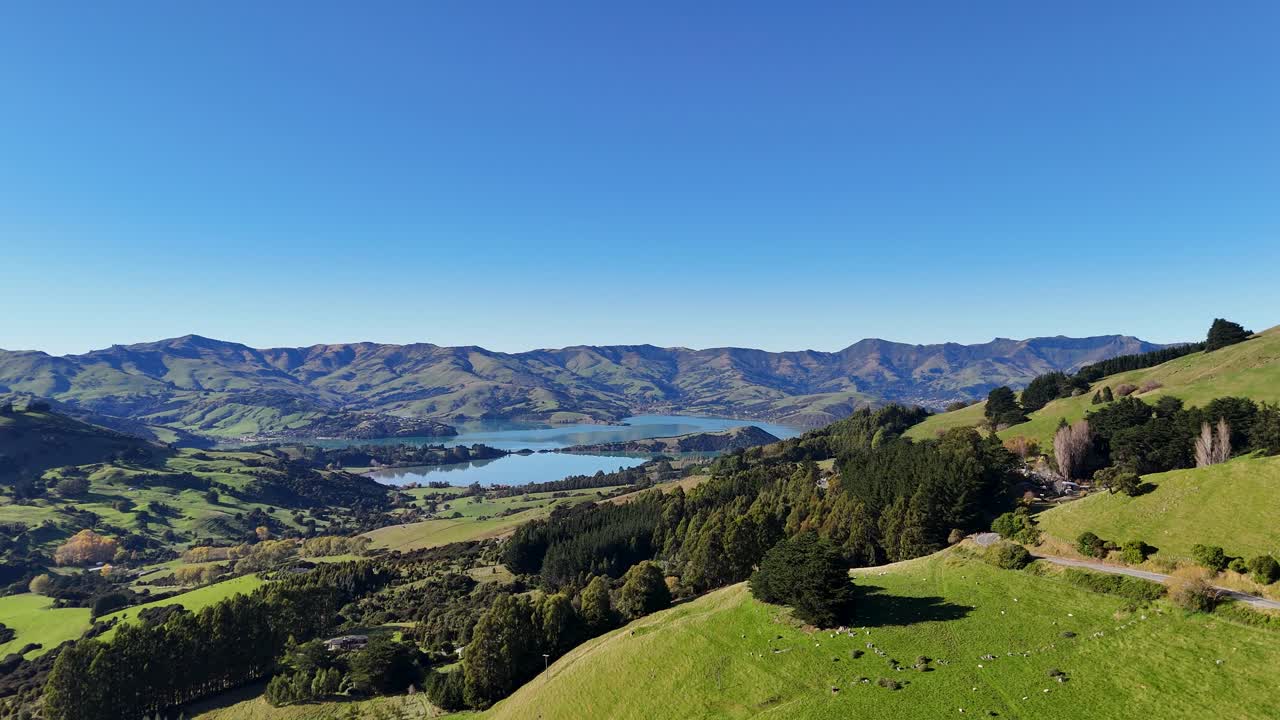 Drone footage captures rolling hills, lush greenery, and a serene lake under clear blue skies in Akaroa, New Zealand