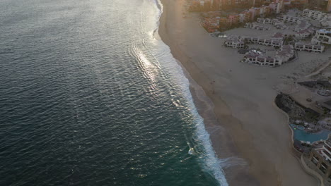 Drone-shot-of-resorts-on-Playa-El-Médano-with-mountains-in-the-distance-in-Cabo-San-Lucas-Mexico,-revealing-and-into-the-sun