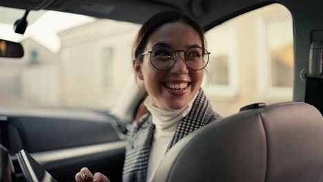 A-happy-brunette-businesswoman-girl-in-round-glasses-and-a-gray-jacket-communicates-with-her-interlocutor-who-is-sitting-in-the-back-seat-while-she-is-sitting-in-the-front-seat-in-a-modern-car-and-looking-at-something-on-her-tablet