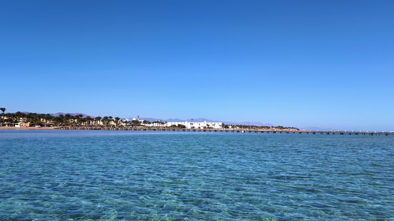 disparo aéreo de un dron volando sobre la superficie del mar con un muelle de madera visible en la distancia en un día soleado