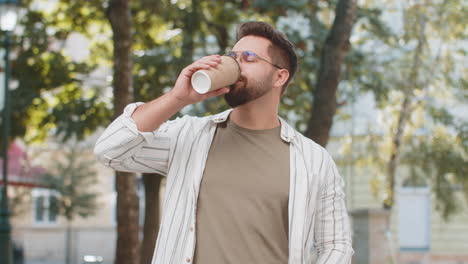 happy caucasian young man tourist in glasses enjoying morning coffee hot drink on city street