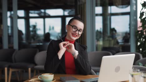 Eine-Wunderschöne-Junge-Frau-Mit-Brille-Und-Zahnspange,-Eine-Freiberuflerin,-Sitzt-Mit-Einem-Laptop-In-Einem-Stilvollen-Restaurant,-Trägt-Business-Kleidung-Und-Eine-Brille,-Lächelt,-Winkt-Zur-Begrüßung-Und-Spricht-In-Einem-Videoanruf