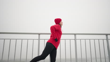 madre de mediana edad haciendo ejercicio al aire libre durante el invierno, sosteniendo una barandilla de hierro para el equilibrio con la mano en la cintura, estirando la pierna hacia adelante y hacia atrás contra la atmósfera brumosa y los postes de luz de fondo