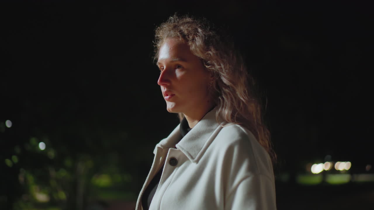 Side view of tired woman in white coat walking alone at night with distant light filtering through trees, captured in low lighting with soft shadows and a peaceful yet moody nighttime atmosphere