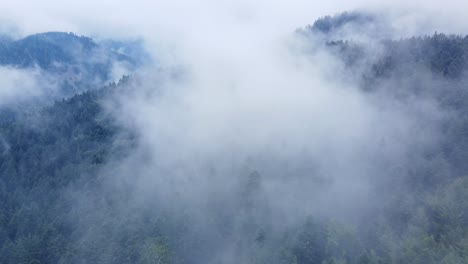 Aerial-view-going-through-big-moody-white-clouds-above-a-dark-mountain-forest,-in-Vosges,-France,-4K