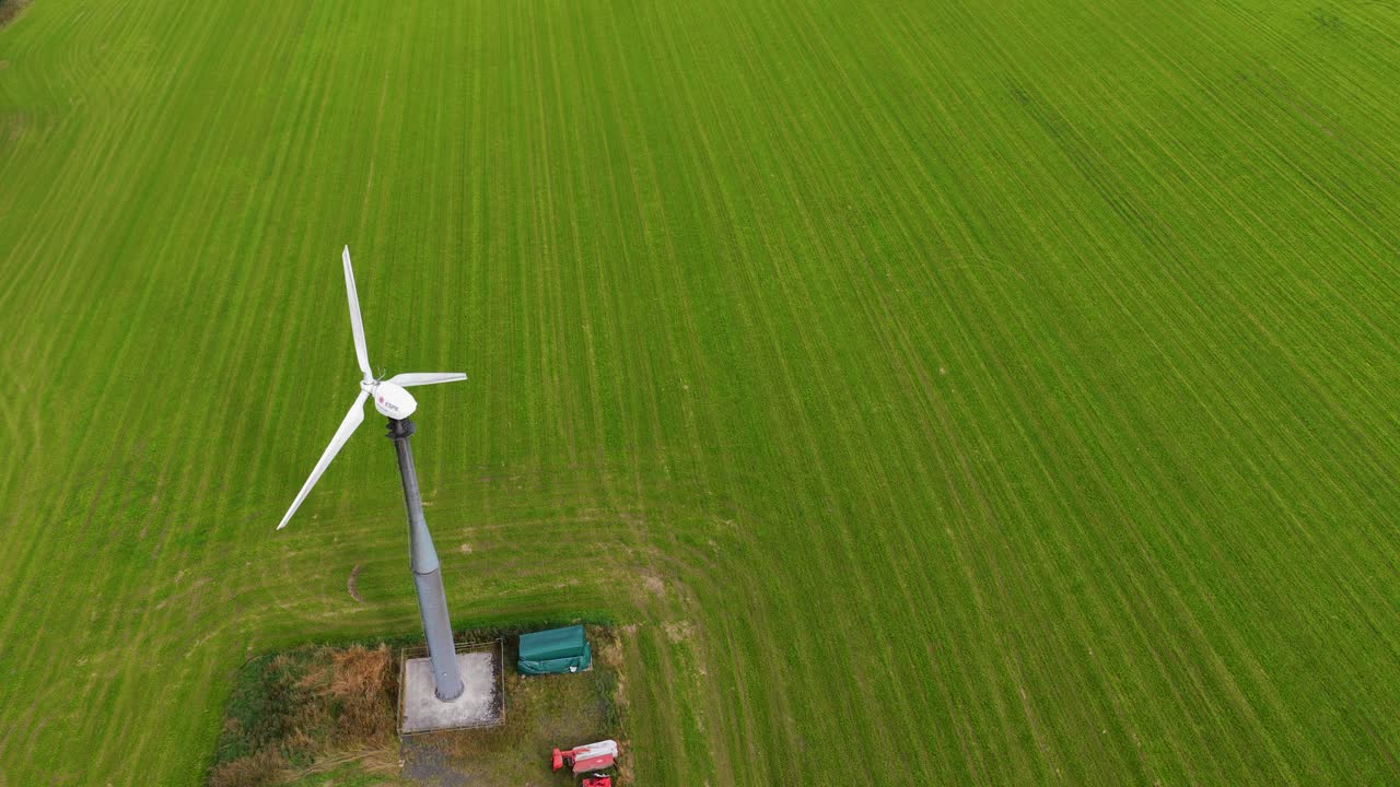 Drone footage captures a single wind turbine spinning steadily above a lush, green agricultural field in Kinross, Scotland, under natural daylight