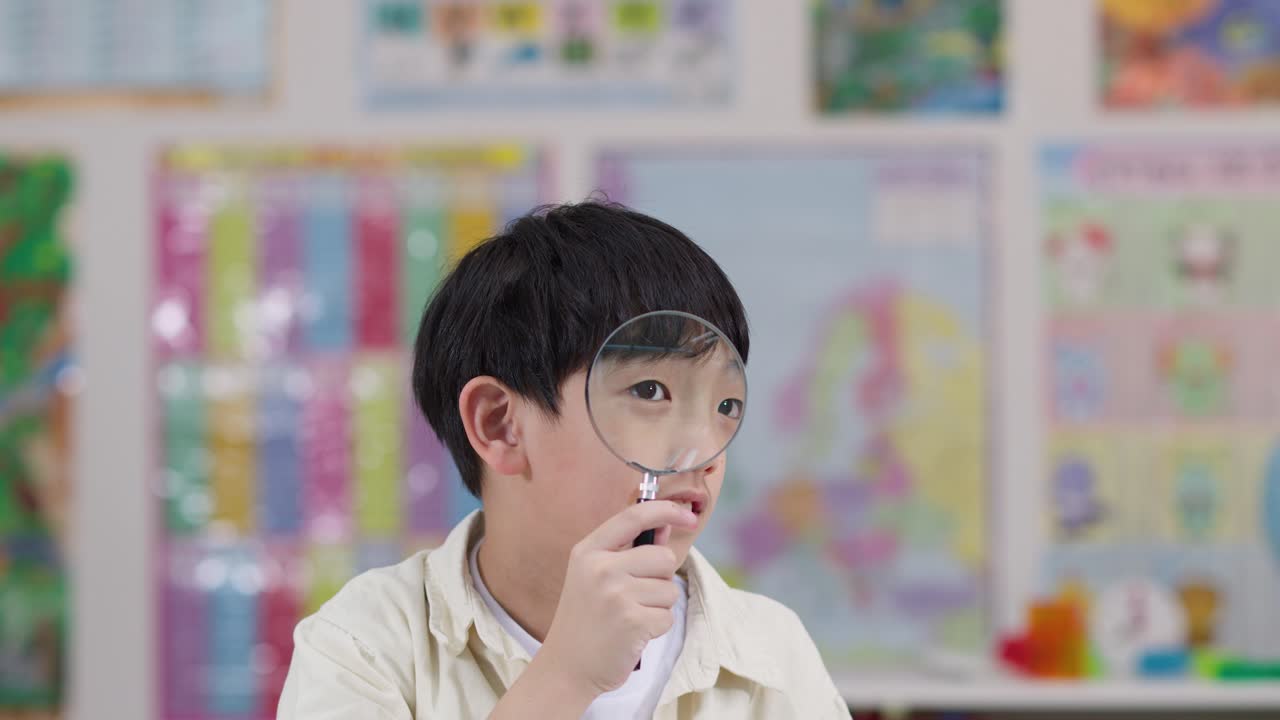 เด็กชายตัวน้อยกำลังสำรวจสิ่งรอบตัวอย่างสนุกสนานด้วยแว่นขยายในห้องเรียนที่สว่างไสว ถ่ายแบบโคลสอัพ