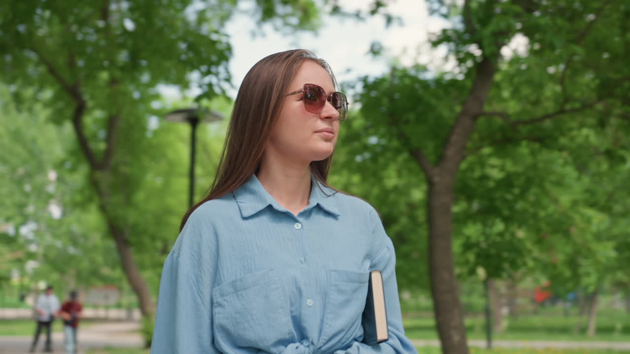 Donna caucasica con dei libri in mano sotto un albero, occhiali da sole, un leggero sorriso, pausa di lettura nel campus, rilassata sicurezza, area con panchina del parco illuminata dal sole, camicia di jeans casual, connubio tra studio e tempo libero