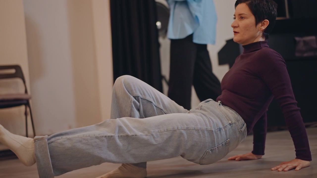 Mature white dancer doing floorwork in studio, concentrated expression with controlled leg extension and core engagement, casual jeans and sweater contrast with formal practice, calm training