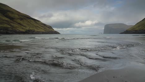 close up of flowing water of sea reaching shore of tjörnuvik beach - risin og kellingin sea stacks in background during cloudy day on streymoy island, faroe islands