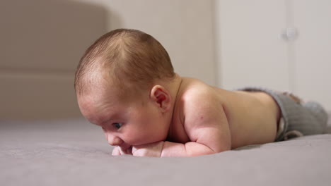 infant lying on the bed on his belly. sweet boy chewing his tiny fists. little kid lying on the bed in a light room close up.