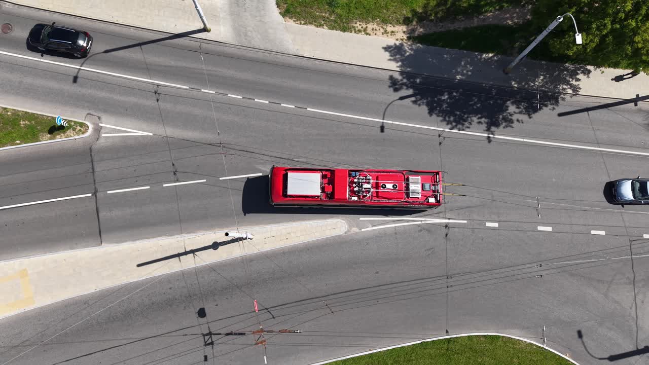 Top-down drone shot of a red trolleybus crossing a wide intersection in Vilnius, following overhead wires and lane markings in bright summer light