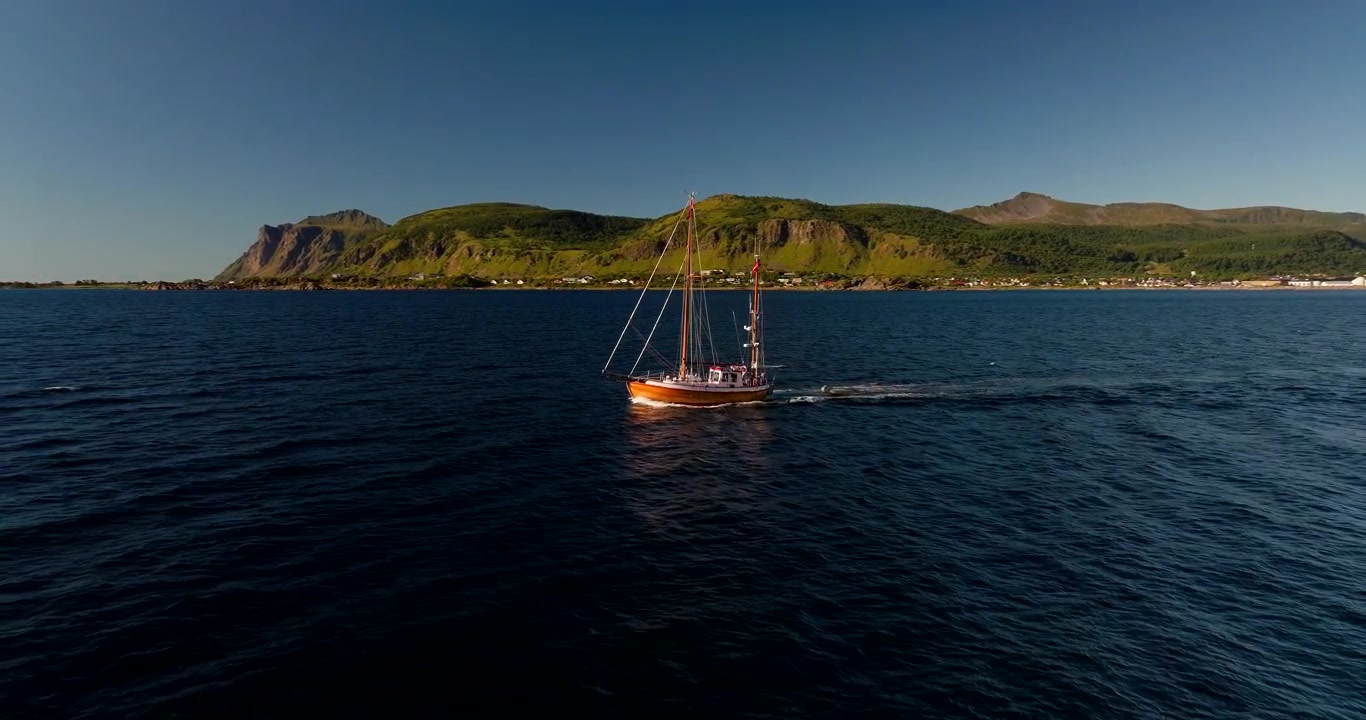 Tradizionale barca a vela in legno che naviga in un fiordo con bellissime montagne nelle Isole Lofoten, Norvegia. Ripresa aerea all'indietro, spazio per il testo.