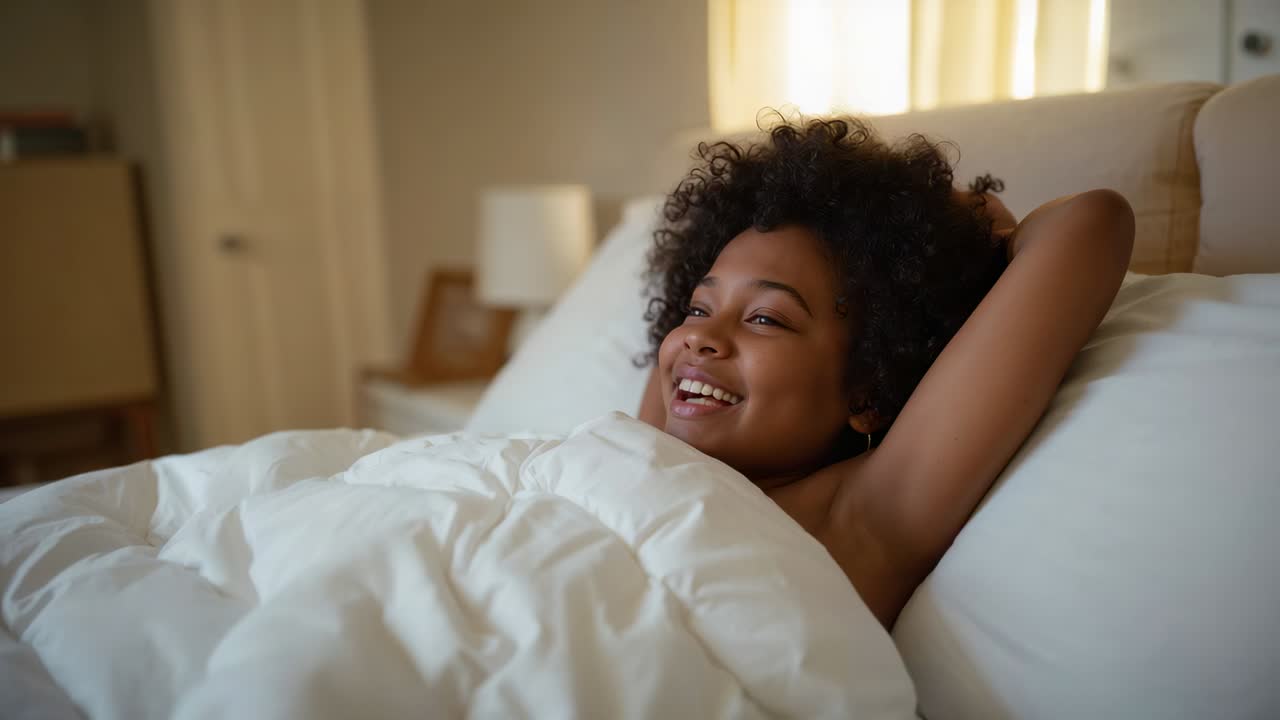 Donna che si sveglia alla calda luce del mattino, sorridendo e stiracchiandosi in camera da letto con piumone e spalle scoperte, appoggiata alla testiera
