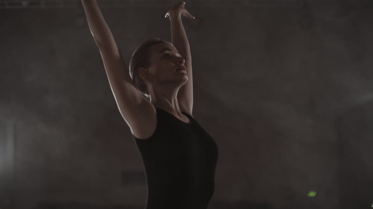 Graceful woman ballerina in a dark dress on a dark stage of the theater in the smoke performs dance moves in slow motion.