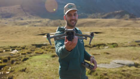 un hombre sonriente en una chaqueta sostiene un dron, listo para volar, con montañas y hierba en el fondo