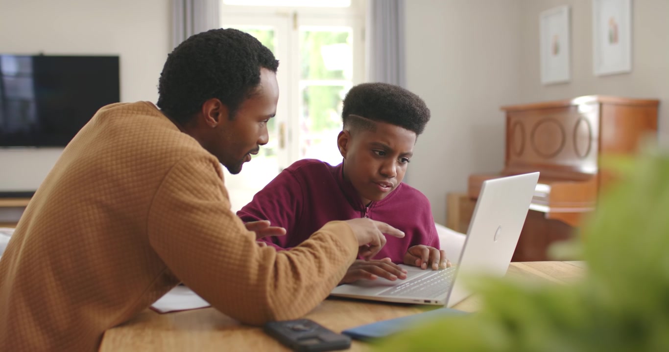 Afrikansk-amerikansk far og tenåringssønn veileder og lærer ved et trebord, den voksne peker på en bærbar PC.
