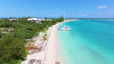 Forward-shot-of-the-beach-with-white-powdered-sand-and-blue-ocean,-people-walking-on-sand,-passing-by-beach-front-villas,-and-big-yachts-anchored-nearby