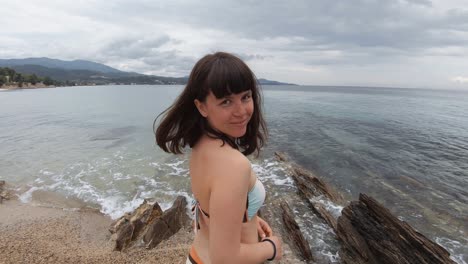 Ein-Junges-Mädchen-Im-Badeanzug-Steht-An-Einem-Felsigen-Strand-In-Griechenland-An-Einem-Teilweise-Bewölkten-Tag-Und-Wendet-Sich-Wieder-Der-Kamera-Zu