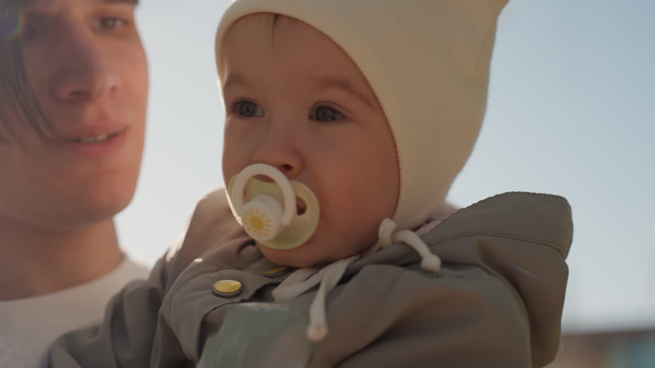 White father holds infant outdoors with pacifier and knit beanie, soft sunlight backlight, gentle gaze and closeup portrait conveying tender parenting and modern family connection in urban setting