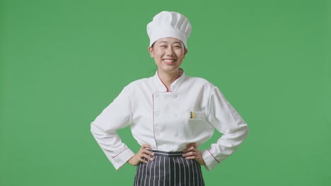 Asian Woman Chef With Arms Akimbo Looking At Camera And Smiling While Standing In The Green Screen Background Studio