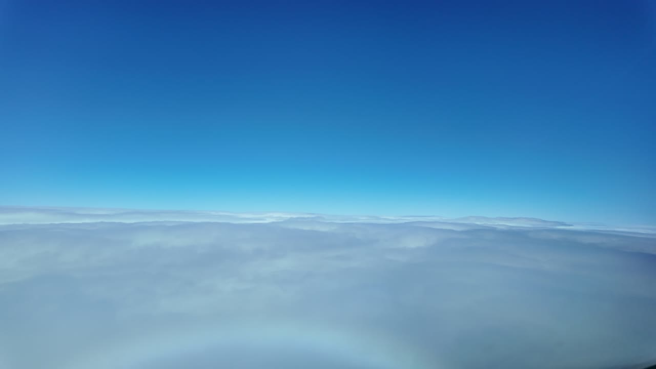Pilot’s eye view flying through a layer of ethereal clouds during descent, with a deep blue sky above, and half a rainbow in the nose of the jet