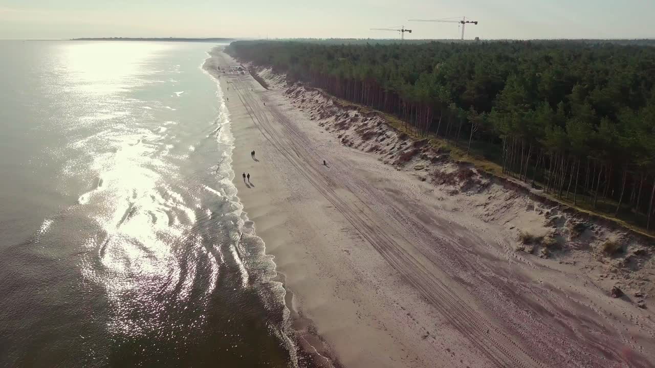 Sunrise on the baltic seashore, sun reflecting on the tranquil waves. Drone footage, few people walking or jogging early in the mornig.