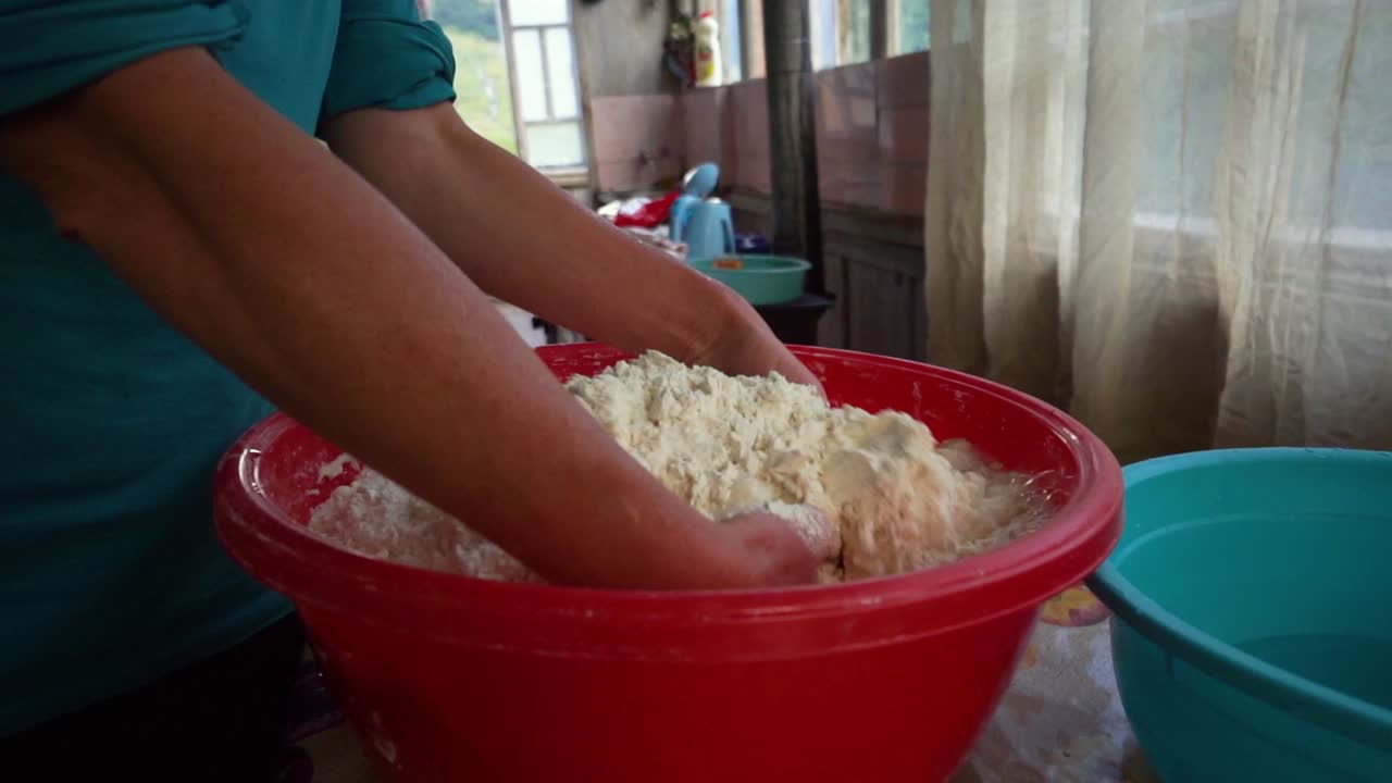 Donna che aggiunge acqua a un impasto per il pane che sta preparando a mani nude in una ciotola di plastica rossa, primo piano, cibo tradizionale georgiano