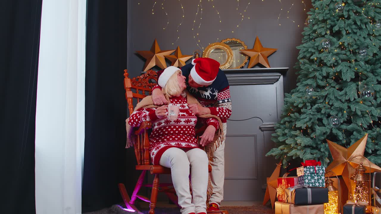 una pareja de ancianos celebra la navidad. el abuelo cubre con una manta a la abuela. la mujer bebe té.