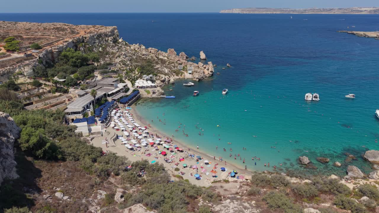 An aerial view of a sandy beach in Malta filled with colorful umbrellas and sunbeds, where tourists enjoy the clear waters. People are swimming, relaxing on the shore, and boats are anchored nearby