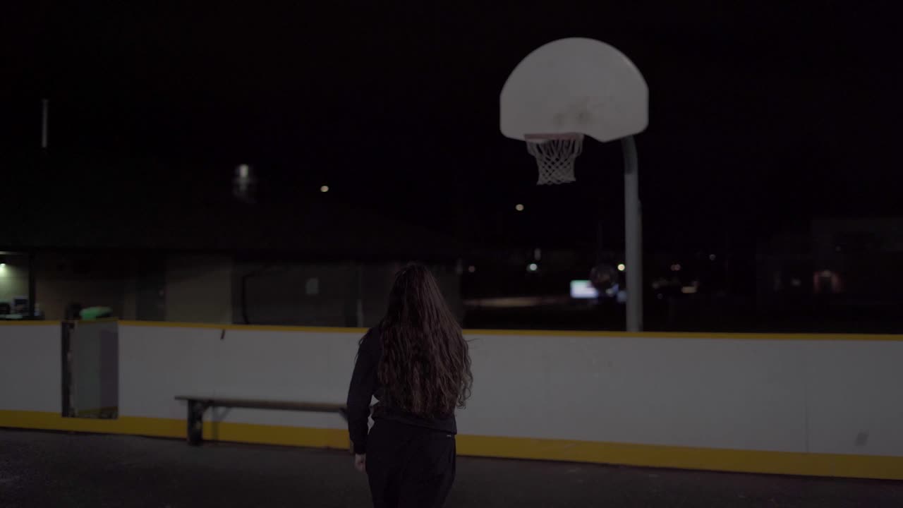 бросает баскетбольный мяч и промахивается на открытой площадке ночью в черном спортивном костюме