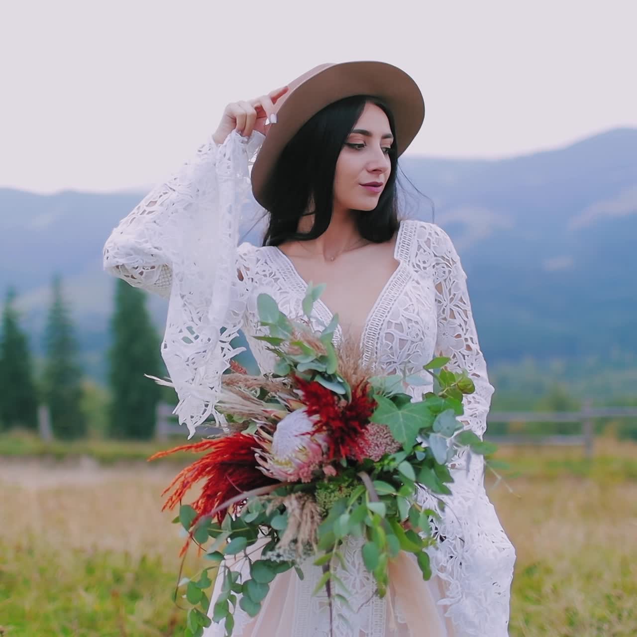 Half-length portrait of a bride in hat outdoors. Beautiful mysterious woman in white dress holds bouquet of flowers on the mountains background.