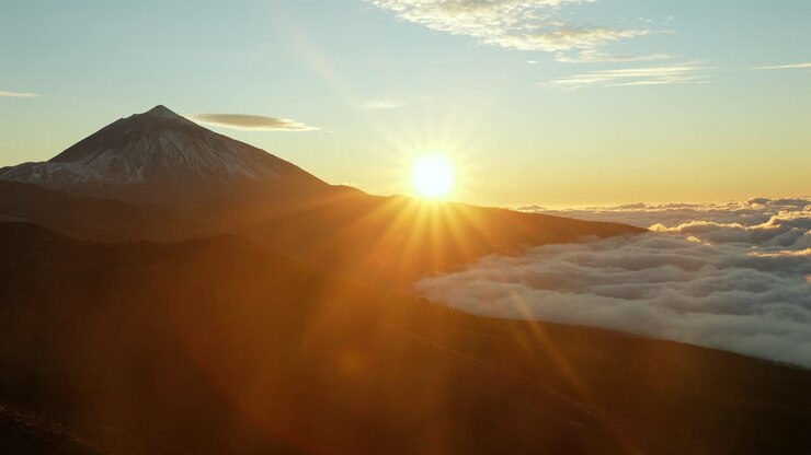 Espectacular escena del sol ascendiendo sobre el famoso volcán Teide en Tenerife. Impresionante paisaje que muestra un pico nevado, cálidos rayos de sol dorados y nubes ondulantes debajo.