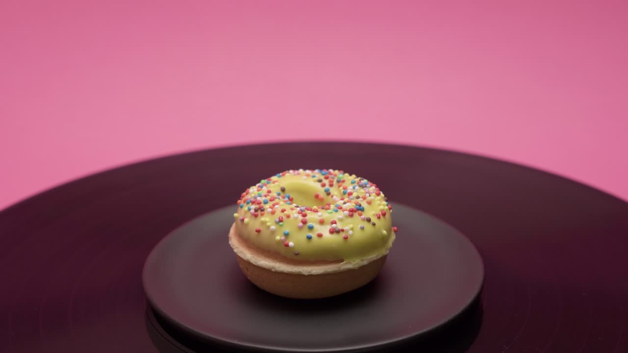 The American donut rolls in circles on a pink and black surface. It is topped with yellow chocolate and garnished with colorful crumbs