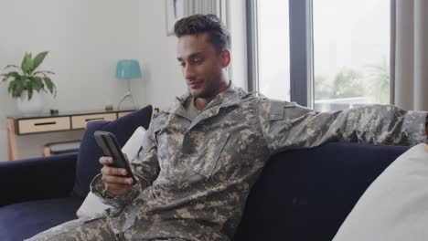 smiling biracial male soldier in uniform sitting on sofa at home using smartphone, slow motion