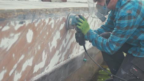 a carpenter sanding paint on the hull of a mahogany planked boat