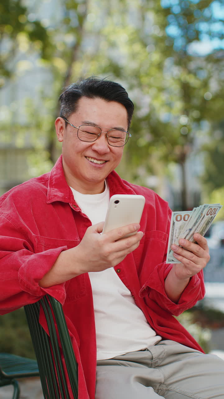 Happy Asian man counting money dollar cash, use smartphone calculator app in urban city street
