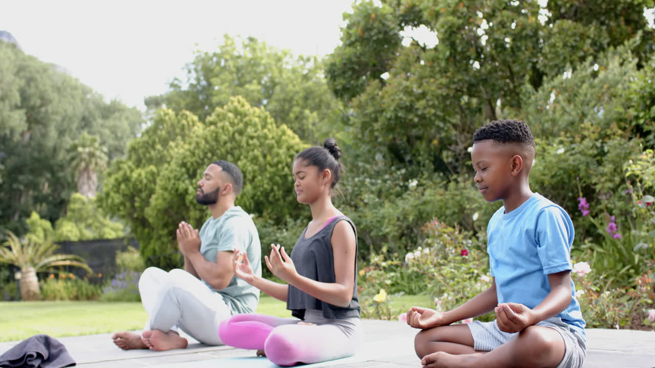 padre afroamericano, hijo e hija practicando meditación de yoga sentados en el jardín, cámara lenta
