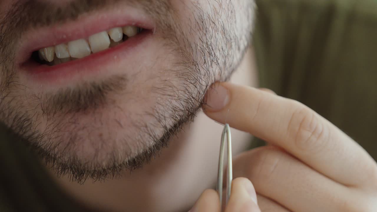 Primo piano di un uomo che indica la zona della barba mentre tiene delle pinzette per rimuovere un pelo incarnito, evidenziando la cura della persona, la cura della pelle e il trattamento di irritazioni minori.