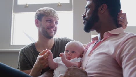 loving male same sex couple cuddling baby daughter on sofa at home together