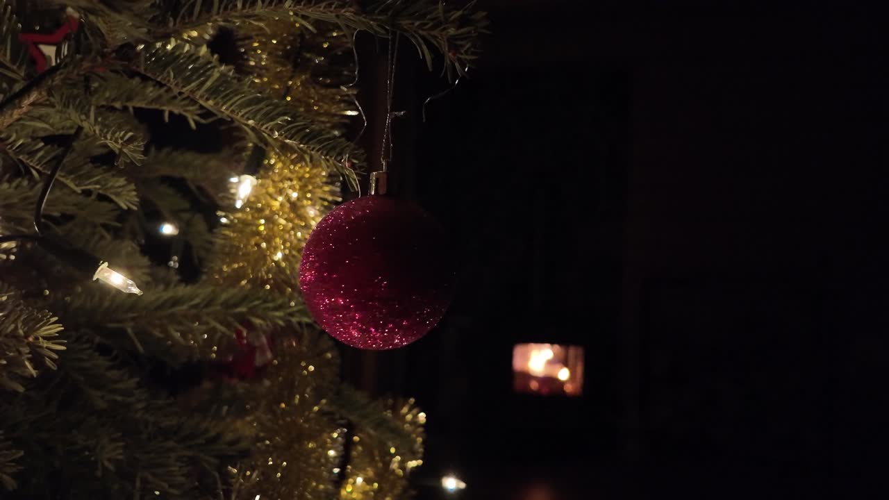 Uma bola vermelha cintilante girando suavemente num pinheiro de Natal decorado, iluminada por luzes festivas quentes numa atmosfera escura e festiva.
