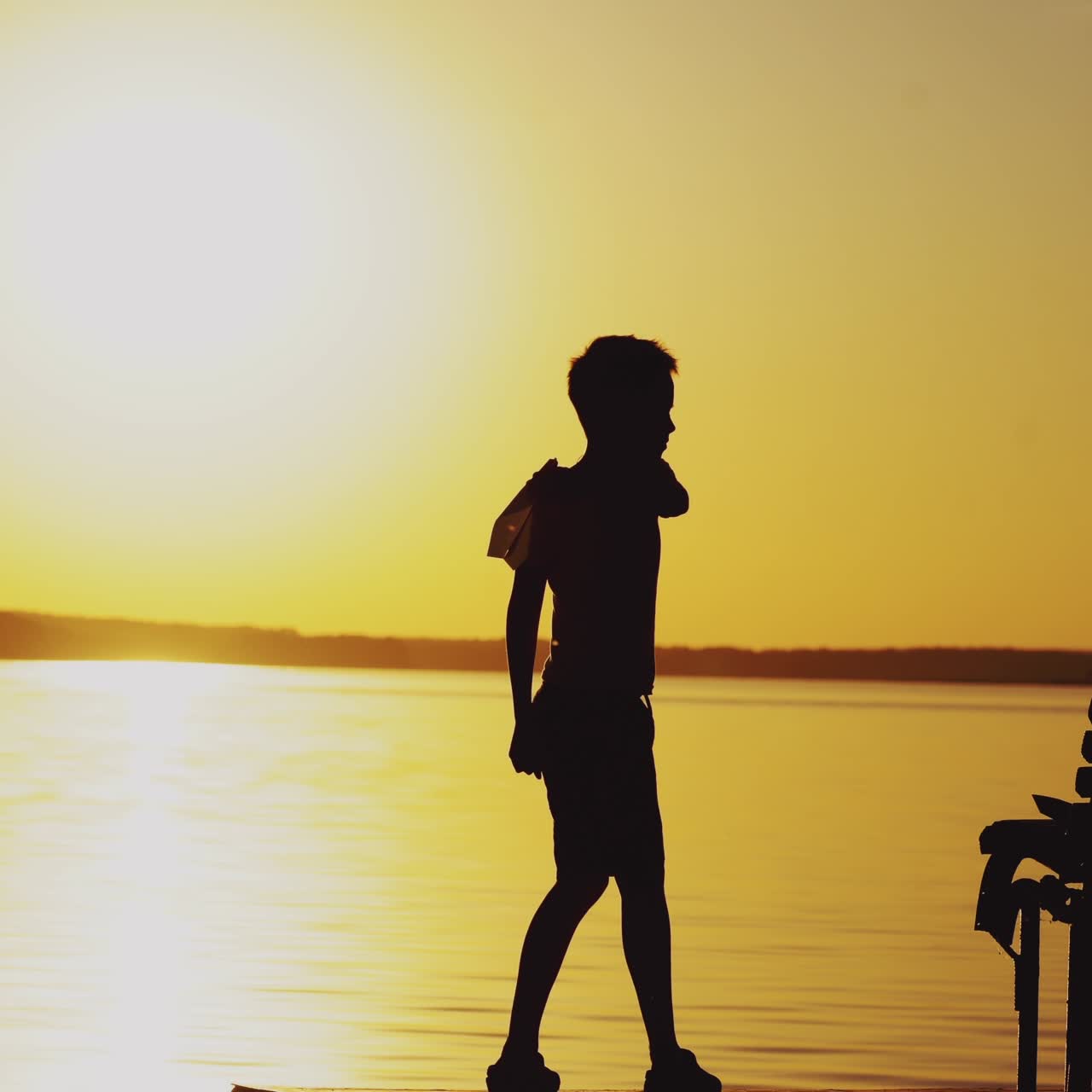 Happy child is playing with a paper airplane. Boy on a wooden bridge on the river in a picturesque setting at sunset.