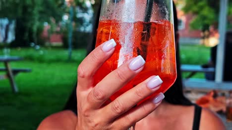 close up of woman with beautiful manicure holding a refreshing aperol spritz cocktail in front of her face while relaxing at an outdoor cafe in summer