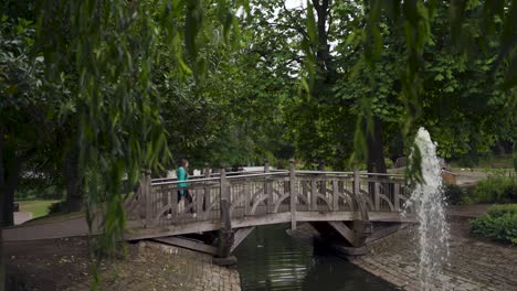 handheld panning shot of young woman in green raincoat walking across wooden bridge in front of fountain in weston park, sheffield, england