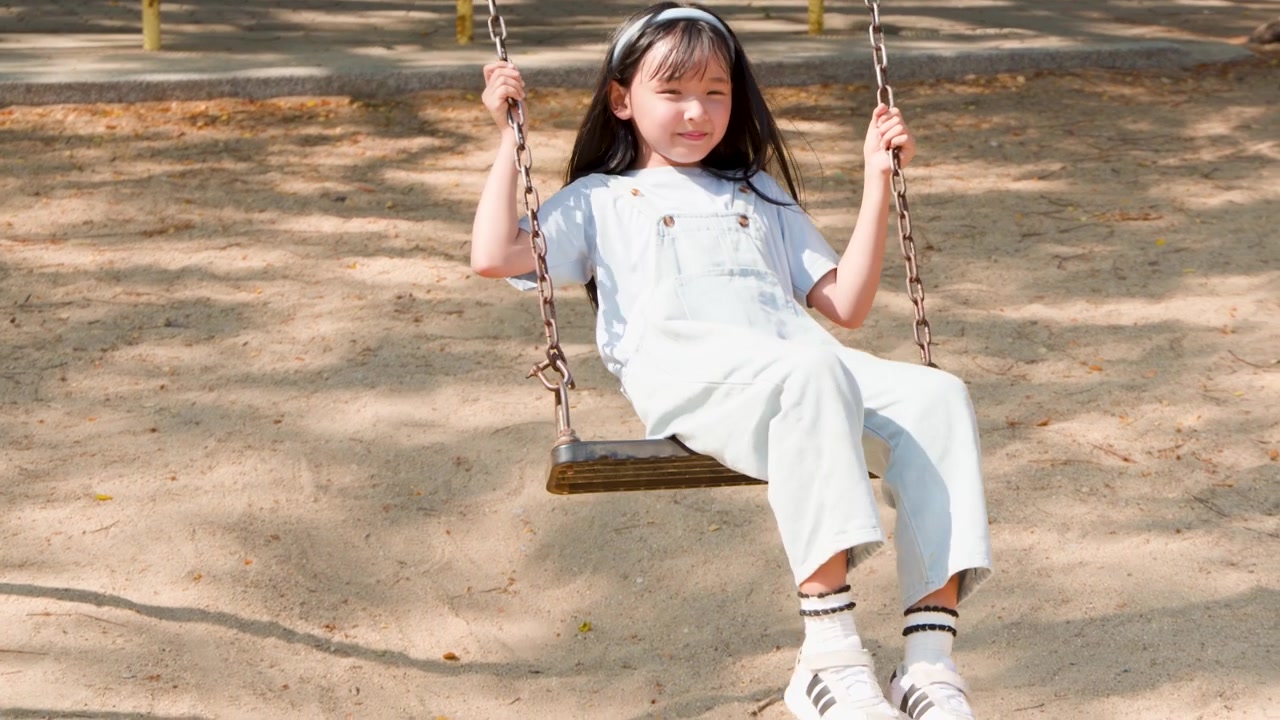 Bambino asiatico in salopette di jeans si diverte sull'altalena in un parco giochi sabbioso a Lumpini Park, Bangkok.