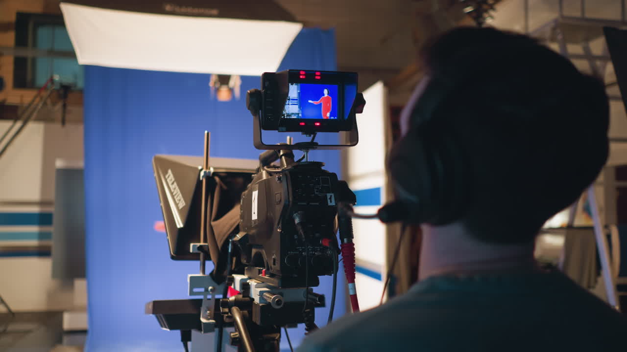 Cameraman wearing headset operating television camera with teleprompter during studio shoot while recording female presenter in red dress on blue screen background under professional lighting setup